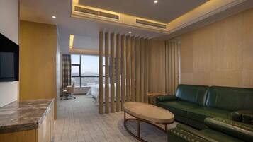 Une chambre d’hôtel moderne avec un canapé en cuir vert, une table basse en bois et une vue sur la ville grâce à de grandes fenêtres.