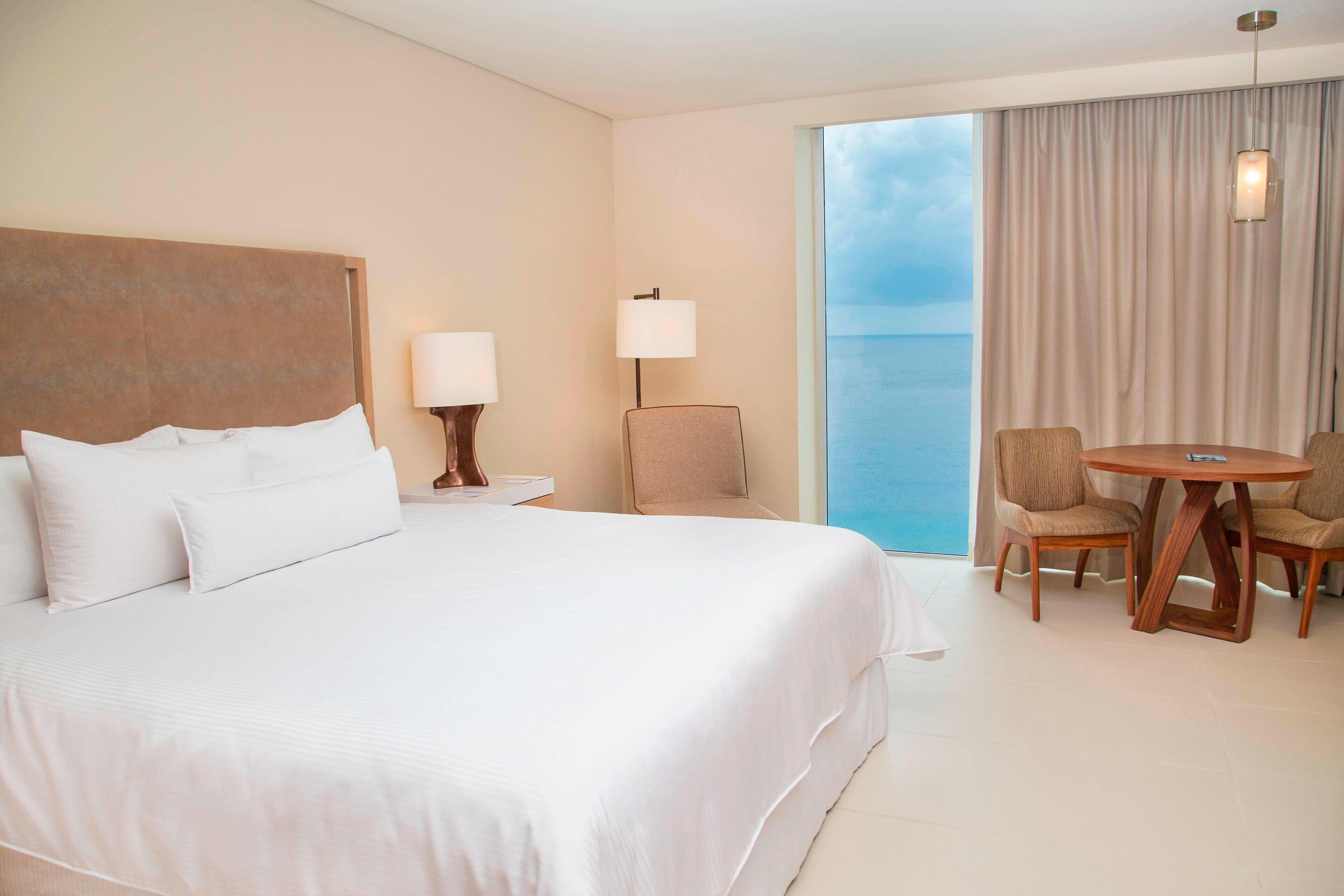 A hotel room with a large bed, a small table with chairs, a lamp, and a view of the sea through a large window.