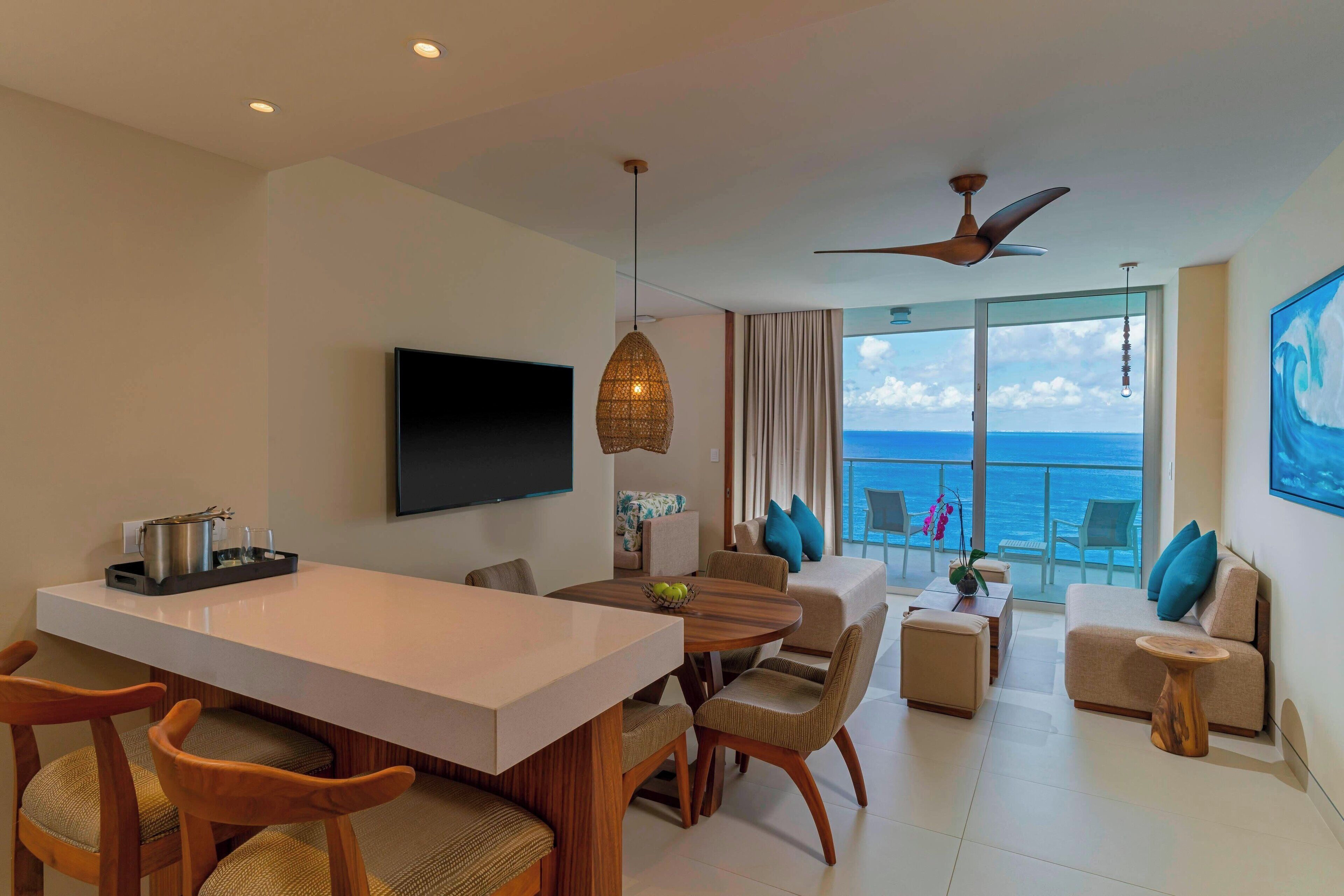 A modern hotel room with a dining area, a sofa set, a TV, and a balcony with a view of the sea.