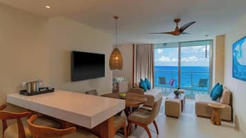 Una habitación de hotel moderna con zona de comedor, un conjunto de sofás, televisión y balcón con vistas al mar.