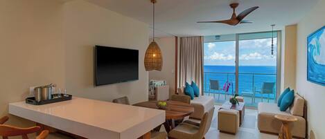 A modern hotel room with a dining area, a sofa set, a TV, and a balcony with a view of the sea.