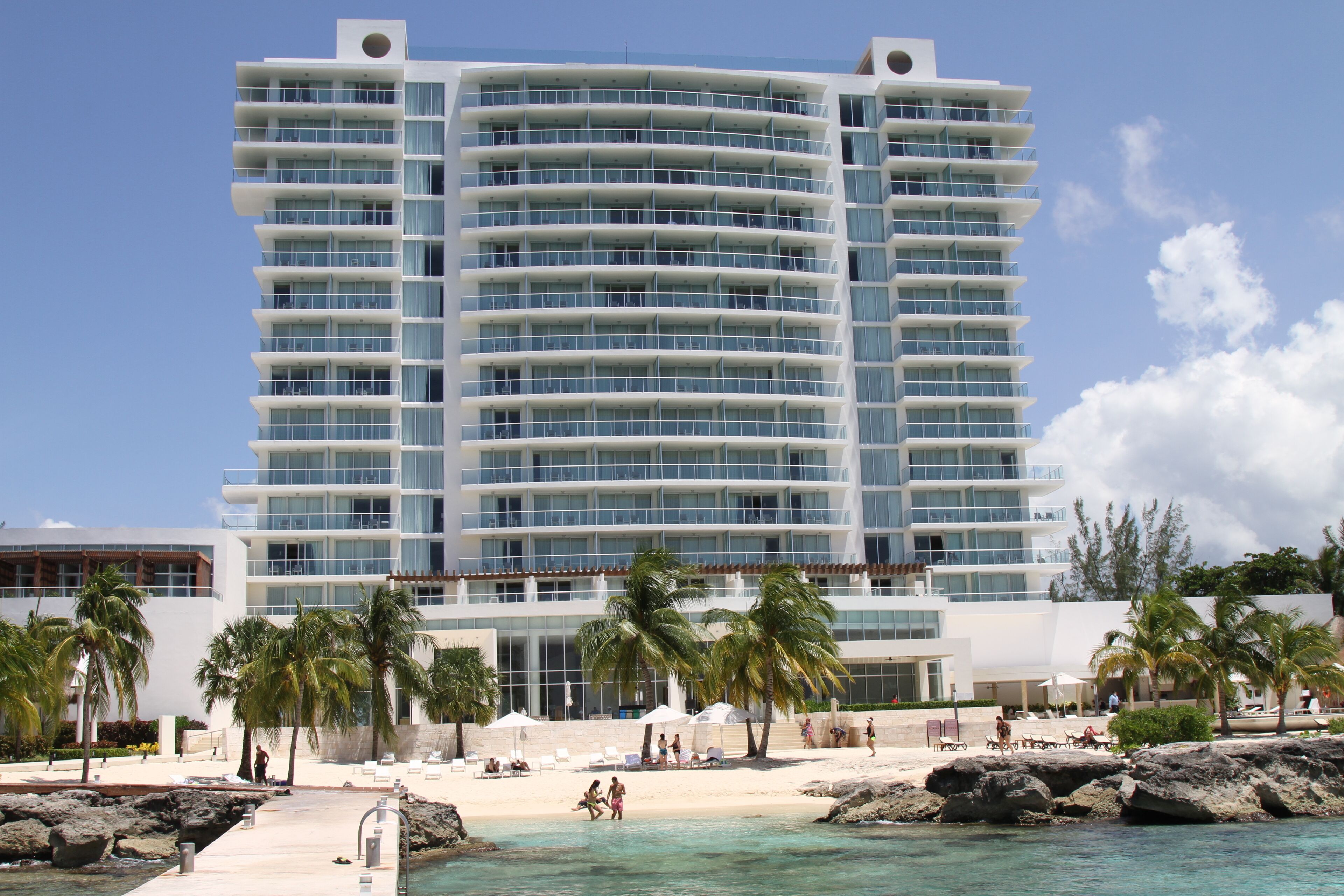 A multi-story hotel with balconies, a beach area with palm trees, and a pool.
