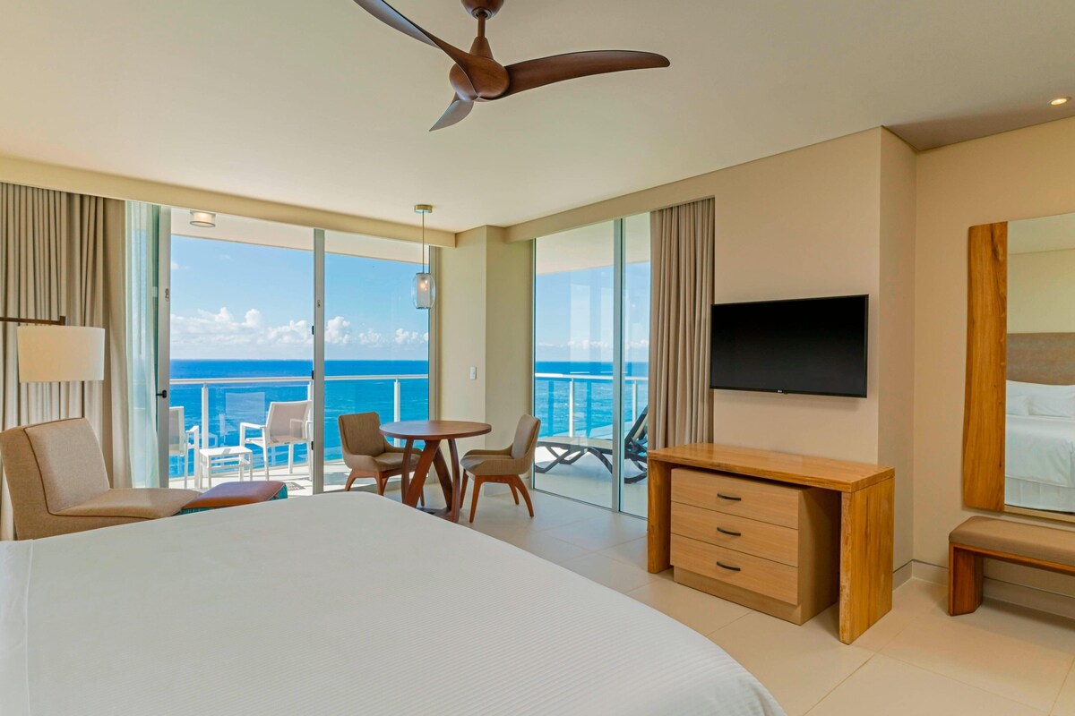 Habitación de hotel con una cama grande, un escritorio, televisor y balcón con vistas al océano.