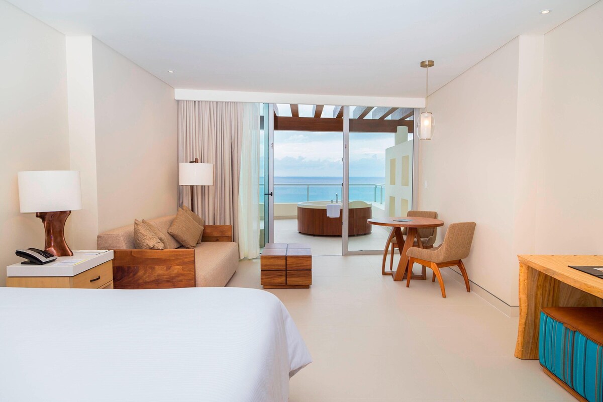 Habitación de hotel con cama, escritorio, sofá y zona de comedor. Se ve el mar a través de las puertas de vidrio.