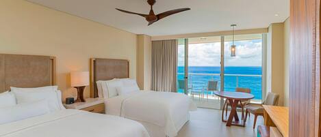 A hotel room with two beds, a ceiling fan, a balcony with a table and chairs, and a view of the ocean.