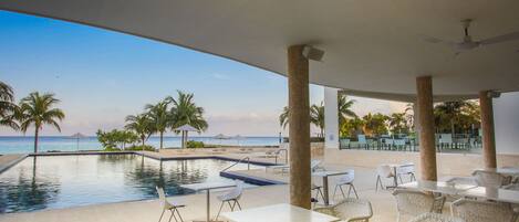 An outdoor pool area with lounge chairs and tables, a clear view of the ocean, and a ceiling fan.