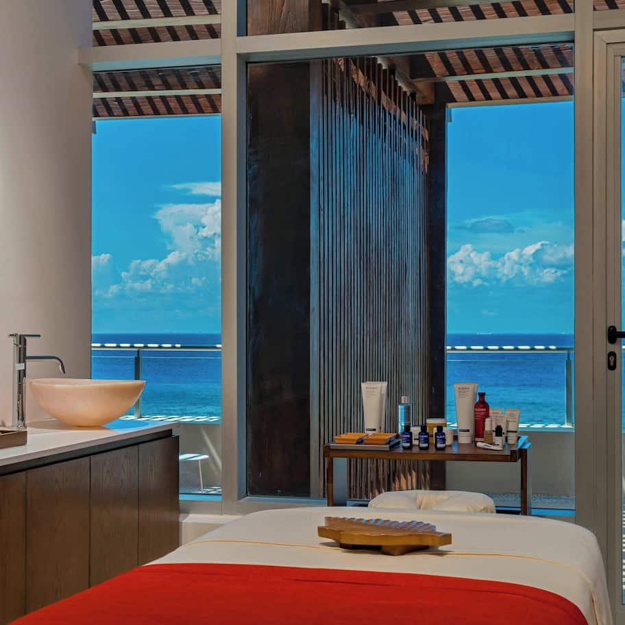 A modern hotel room with a large window overlooking the sea, a bed with a red blanket, a vanity with a large mirror, and a small table with toiletries.