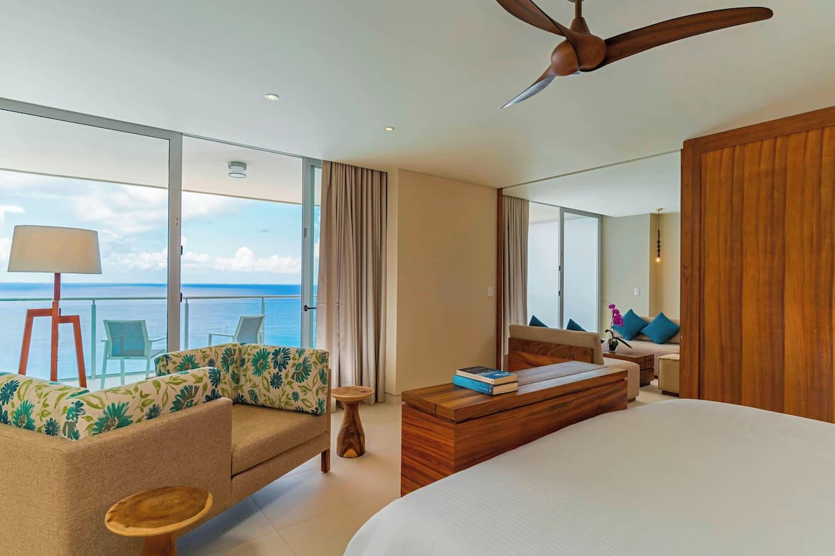 Habitación de hotel moderna con una cama grande, un sofá con cojines estampados, una mesa de centro de madera y vistas al océano.