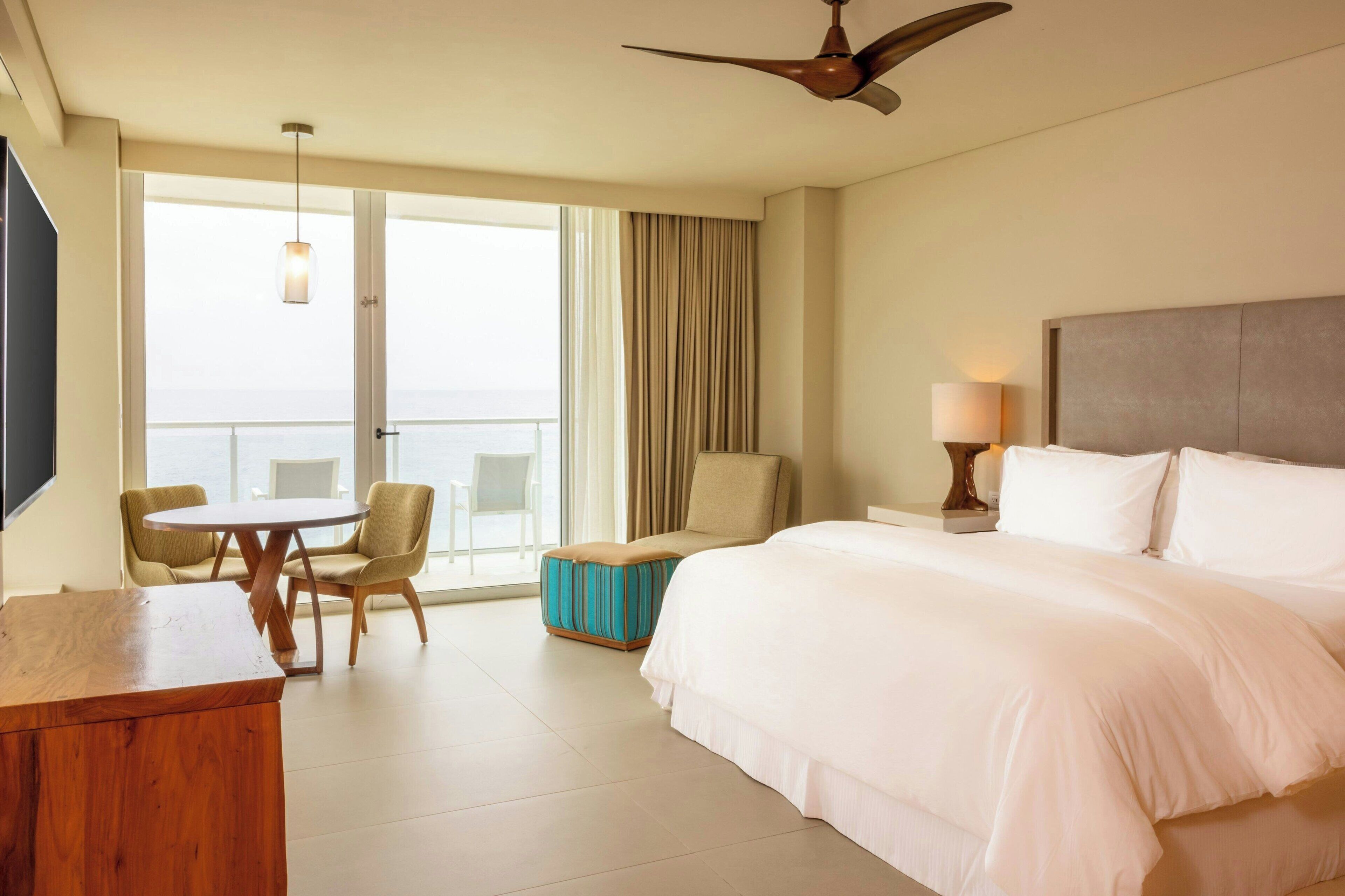 A hotel room with a large bed, a desk, a chair, a small table, and a view of the ocean through the window.