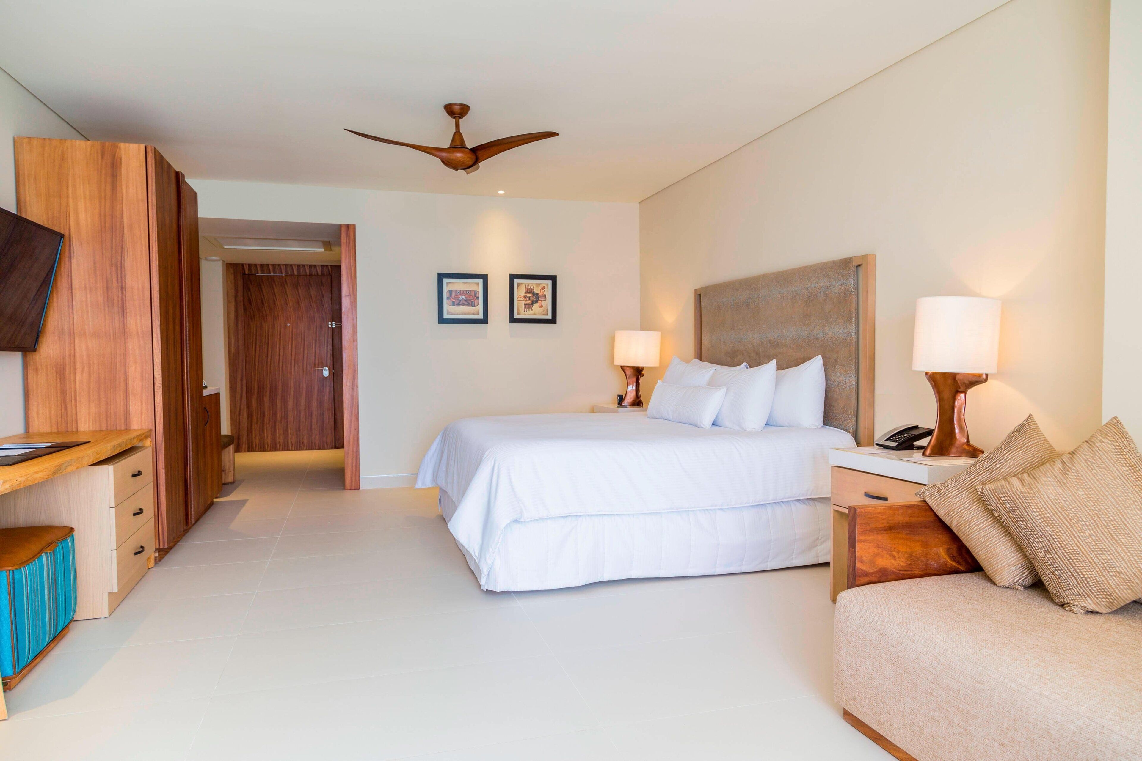 A hotel room with a bed, a desk, a chair, a TV, a ceiling fan, and two wall-mounted lamps.