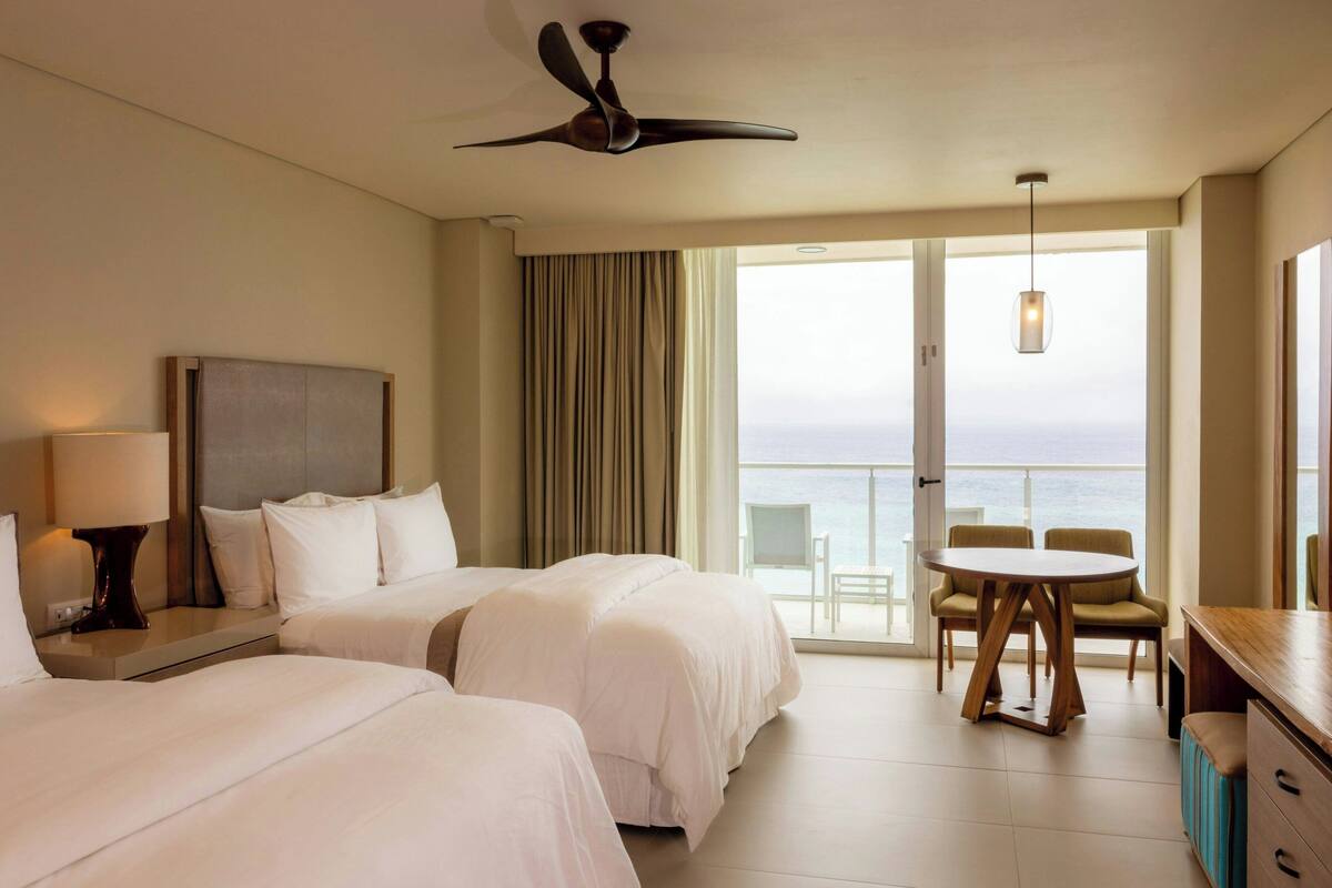 Habitación de hotel con dos camas, un ventilador de techo, una mesa con sillas y vista al mar.