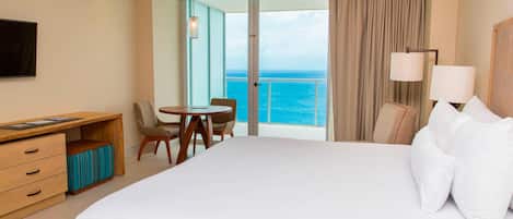 A hotel room with a large bed, a desk with a chair, a small table with a chair, and a view of the ocean through a glass door.