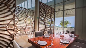 Uma sala de jantar com uma mesa de madeira posta para quatro pessoas, janelas amplas e vista para o exterior.