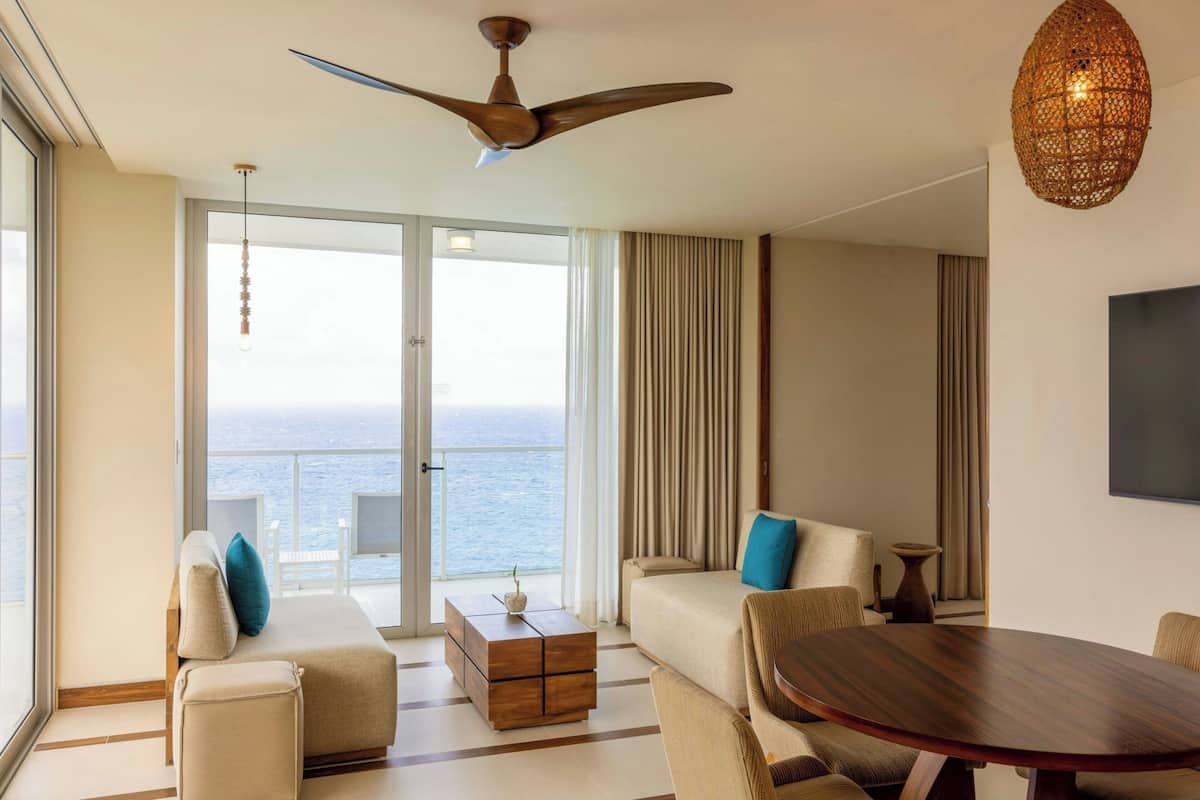 Habitación de hotel moderna con balcón, ventilador de techo, mesa redonda de madera y vista al mar.
