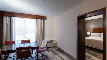 Quarto de hotel com cama, escrivaninha com duas cadeiras, janela grande com cortinas e ar-condicionado fixado na parede.