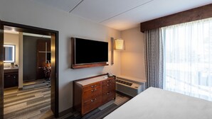 Een hotelkamer met een groot bed, een flatscreen televisie aan de muur, een houten dressoir, een bureau met stoel, en een groot raam met gordijnen.