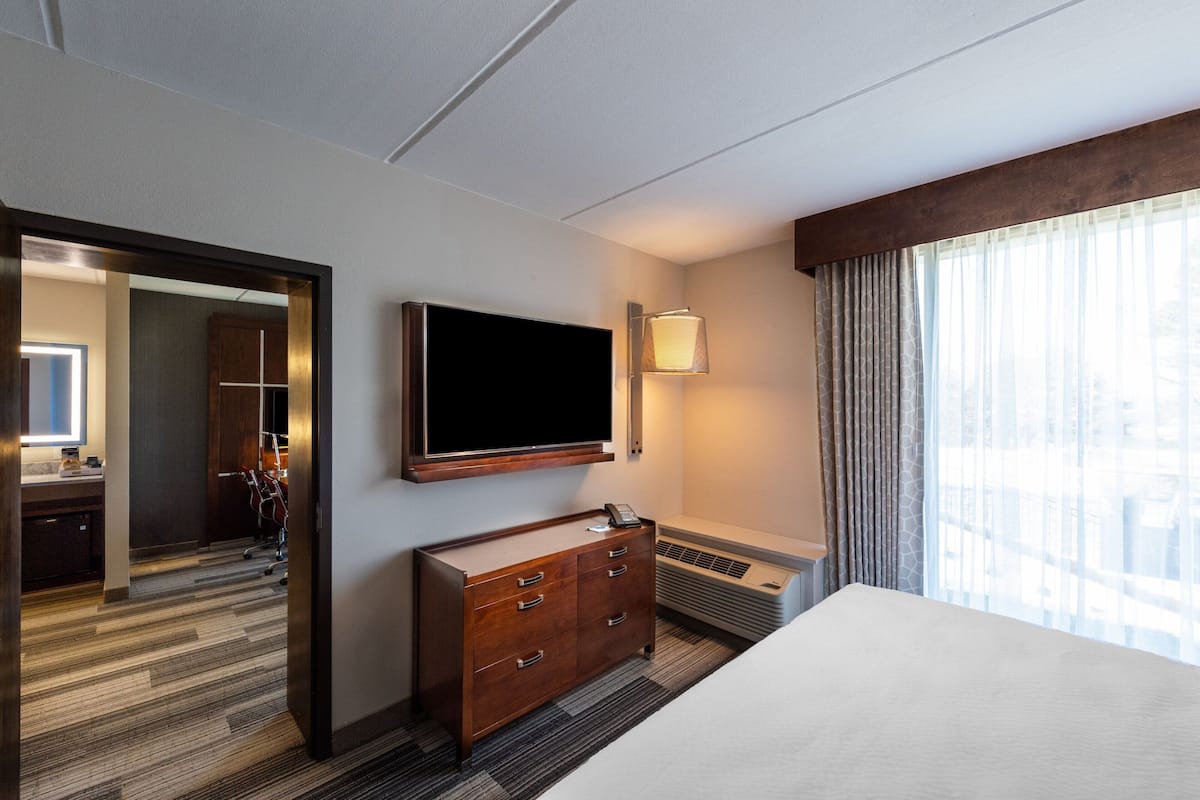 Une chambre d’hôtel avec un grand lit, un téléviseur à écran plat fixé au mur, une commode en bois, un bureau avec une chaise et une grande fenêtre avec des rideaux.