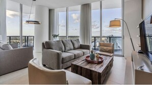Una sala de estar moderna con un sofá gris, una mesa de centro de madera y un televisor. Amplias ventanas ofrecen vista a la ciudad.