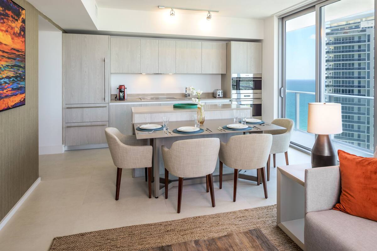 A modern kitchen with light wood cabinets, a dining table with chairs, and a view of the ocean through large windows.