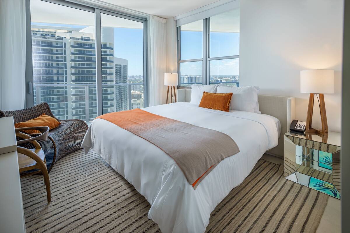 A hotel room with a large bed, a chair, a desk, and a view of the cityscape through large windows.