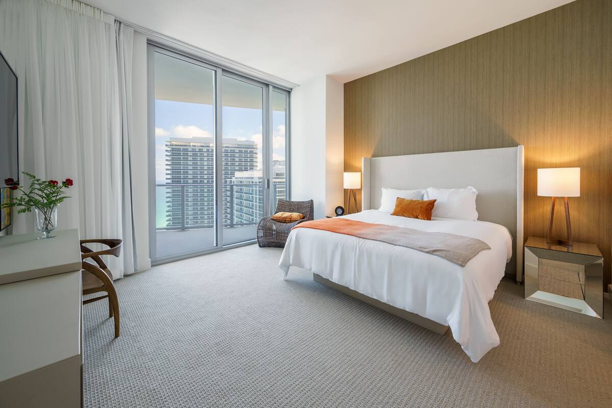A hotel room with a large bed, a desk, a chair, and a view of the cityscape.