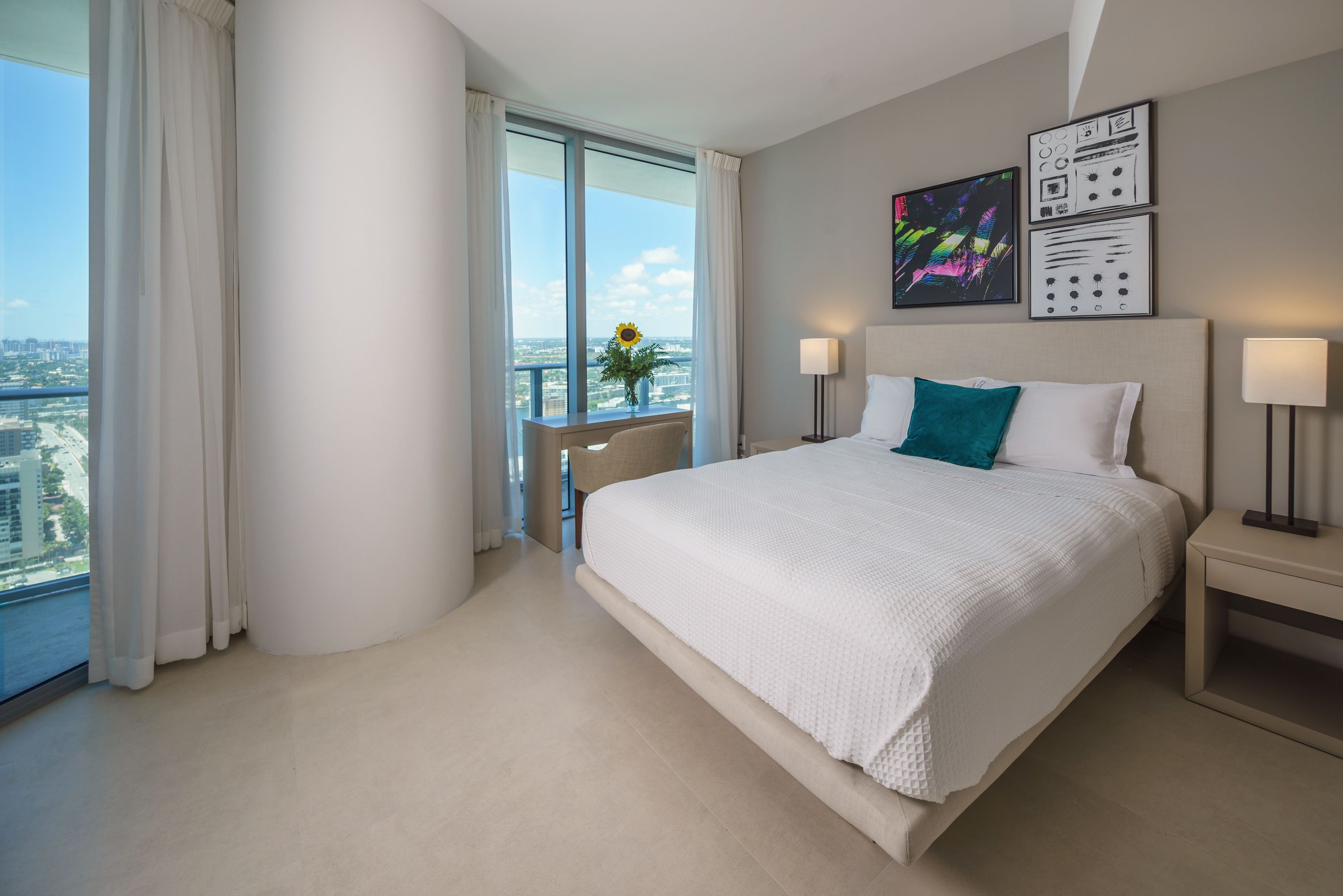 Ein modernes Hotelzimmer mit einem großen Bett, einem kleinen Tisch mit einer Vase und Blick auf die Stadt durch große Fenster.