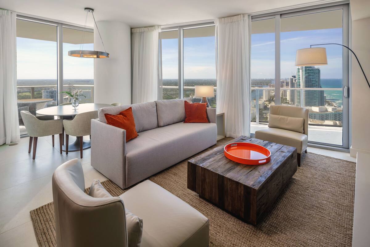 A modern living room with a grey sofa, beige armchairs, a round coffee table, and a dining table with chairs. Large windows offer a view of the city and ocean.