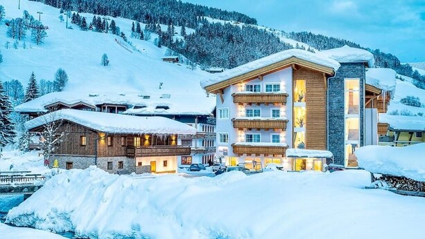 Ein mehrstöckiges Hotel mit Holzfassade und Balkonen, umgeben von schneebedeckten Bergen und einem Fluss.