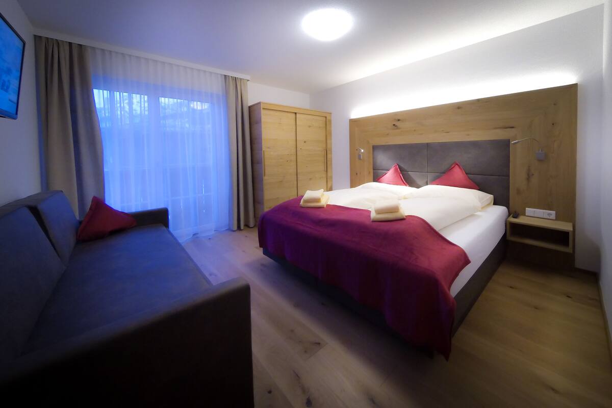 A hotel room with a bed, a sofa, a wooden wardrobe, and a window with curtains.