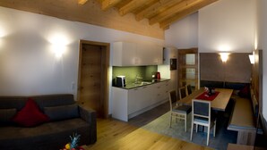 Ein modernes Wohnzimmer mit Holzboden, eine Essecke mit Tisch und Stühlen sowie eine Küche mit weißen Schränken und einer grünen Spritzschutzwand.