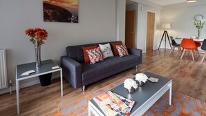 Una sala moderna con un sofá gris, una mesa de centro con revistas, un comedor con mesa y sillas, y un cuadro en la pared.