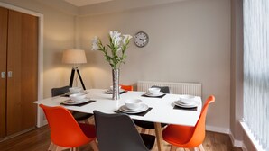 Un comedor con una mesa blanca, sillas anaranjadas y grises, un jarrón con flores y un reloj en la pared.