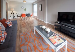 Una sala de estar moderna con un sofá gris, un tapete estampado anaranjado, una mesa de centro con revistas, un televisor y un comedor con una lámpara y sillas.