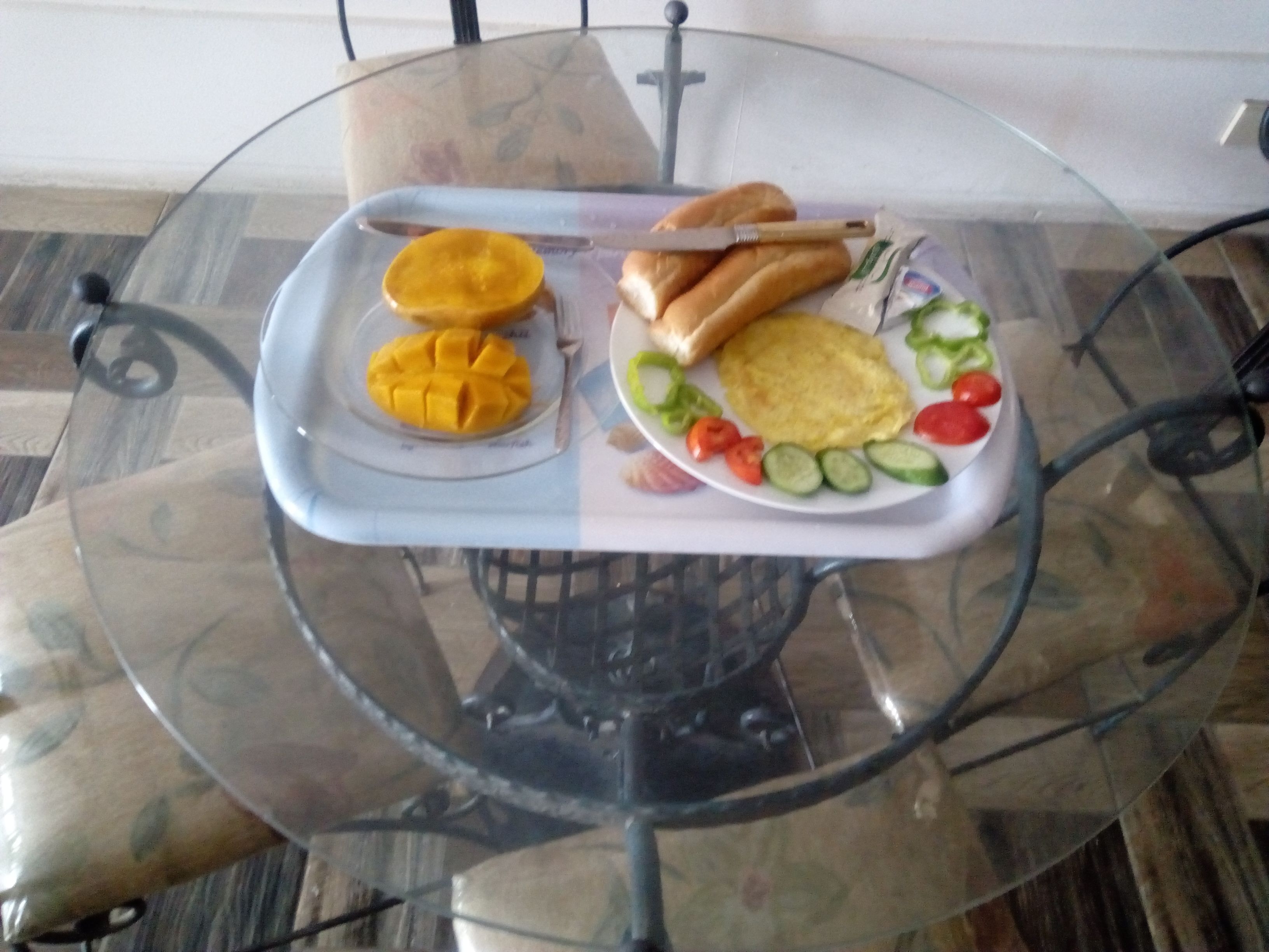 A glass table with a tray containing a mango, two hot dogs, a plate with eggs, tomatoes, cucumbers, and bell peppers.