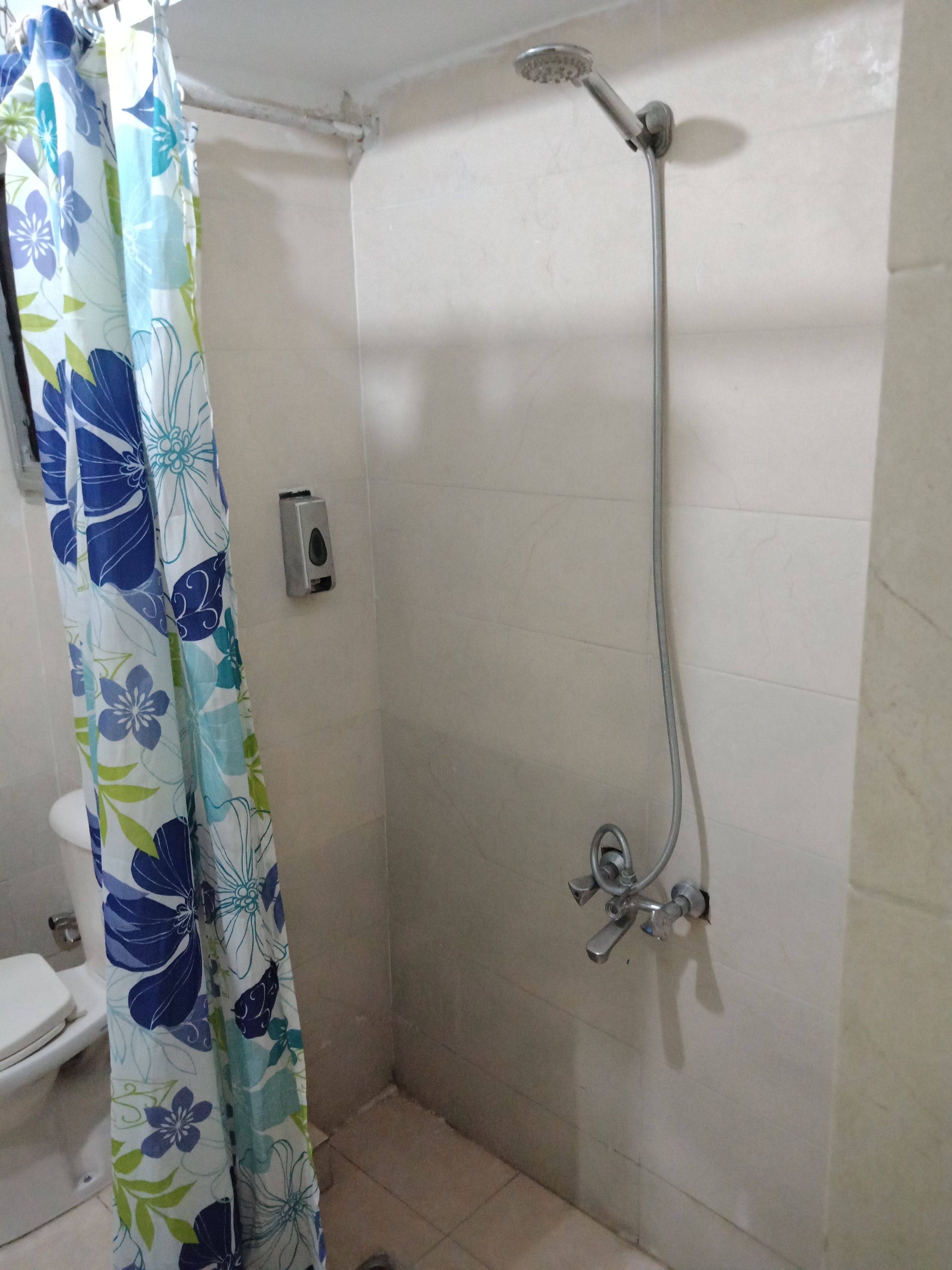 Une douche avec un pommeau de douche amovible, un rideau et des toilettes dans une salle de bain dont les murs sont recouverts de carrelage.