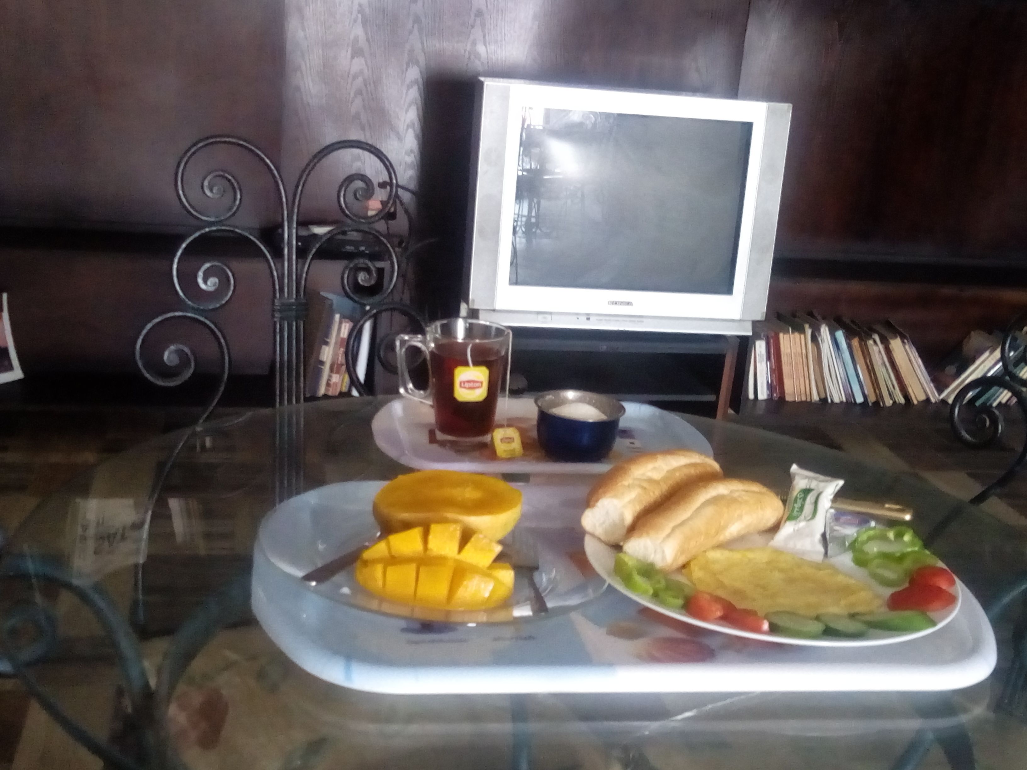 A glass table with a meal consisting of bread, vegetables, and a mango. A cup of tea and a bowl of soup are also visible.