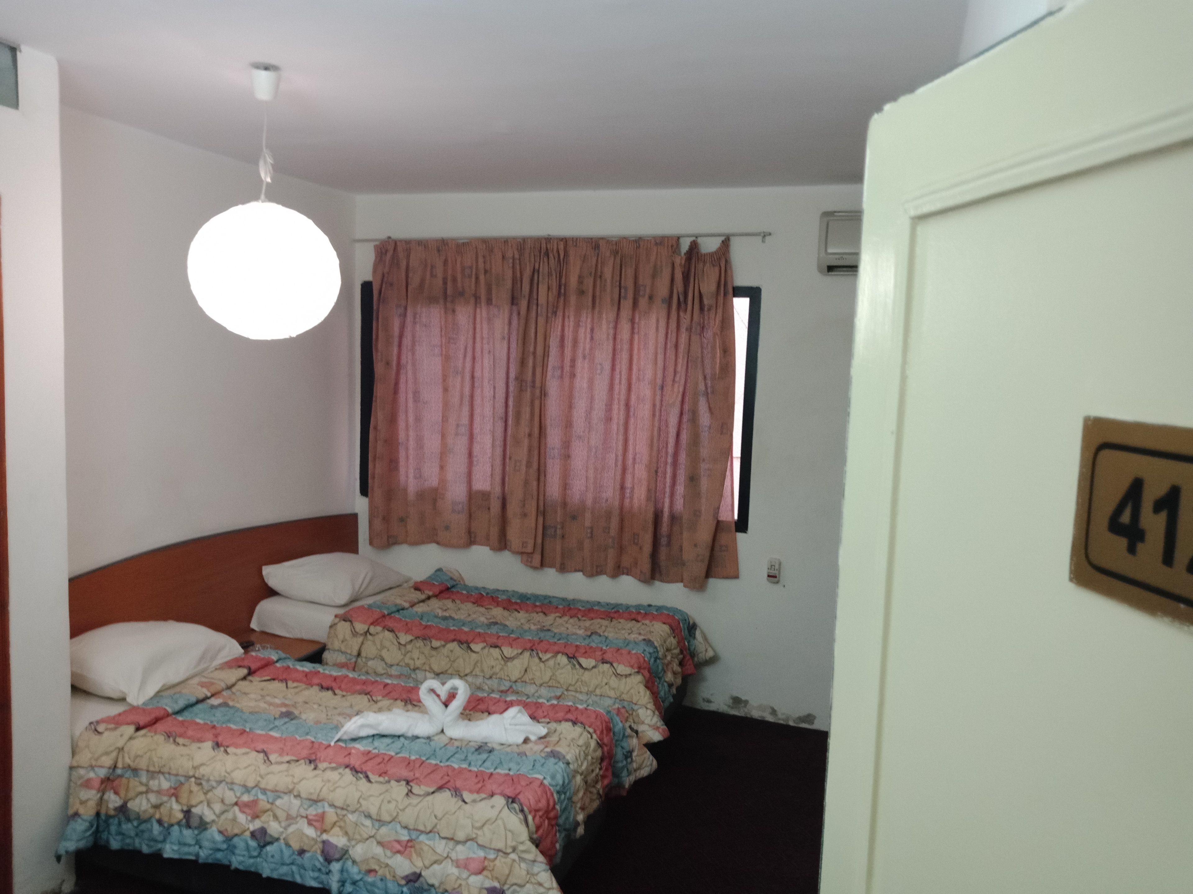 A hotel room with two beds, a window with curtains, a ceiling light, and a door with a number plate.