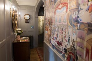 Un couloir avec une horloge, un miroir fixé au mur et un mur décoratif orné d’une fresque.