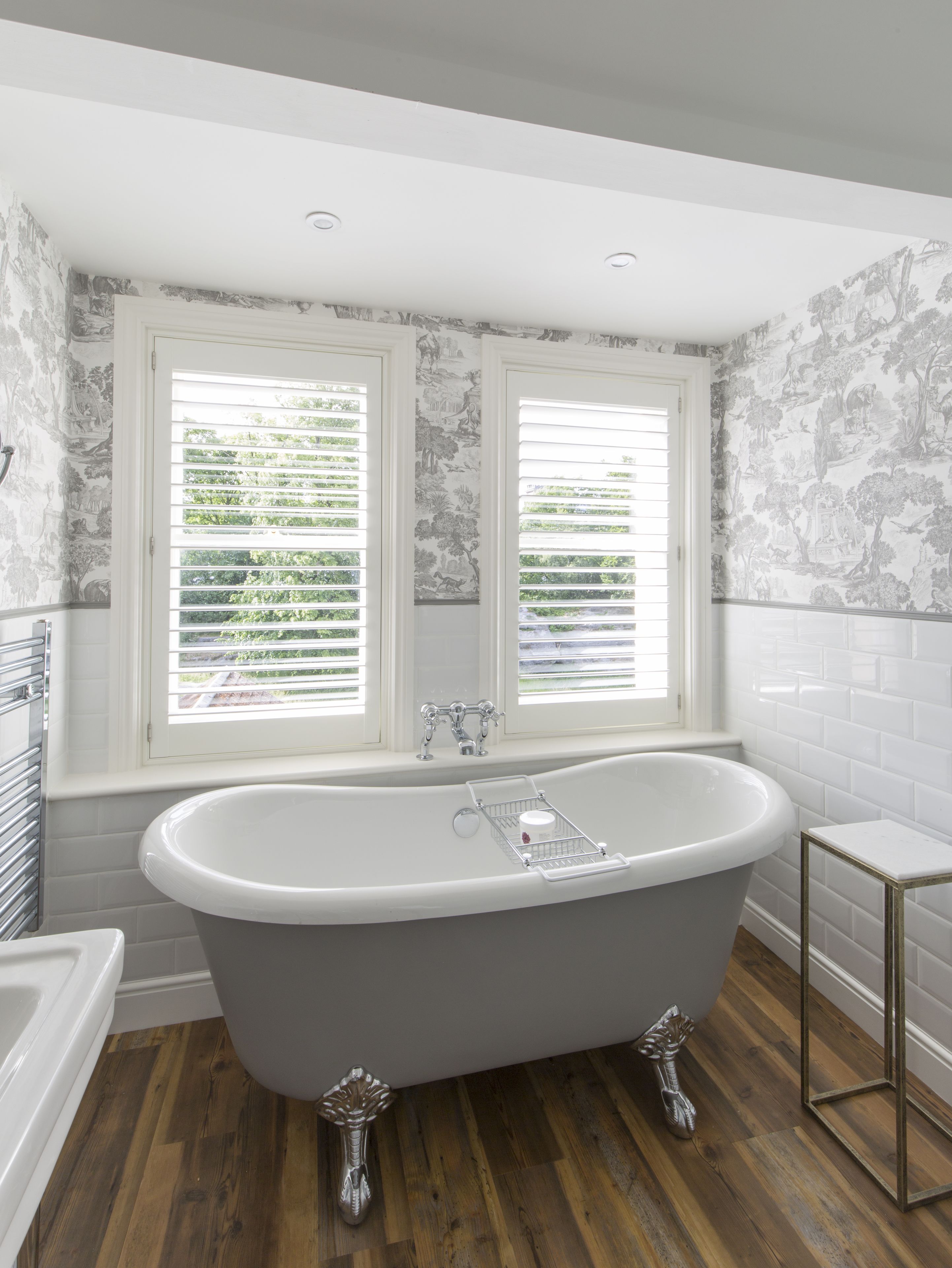 Un baño con una bañera independiente, dos ventanas con contraventanas y una pared de azulejos con estampado floral.