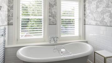 Un baño con una bañera independiente, dos ventanas con contraventanas y una pared de azulejos con estampado floral.