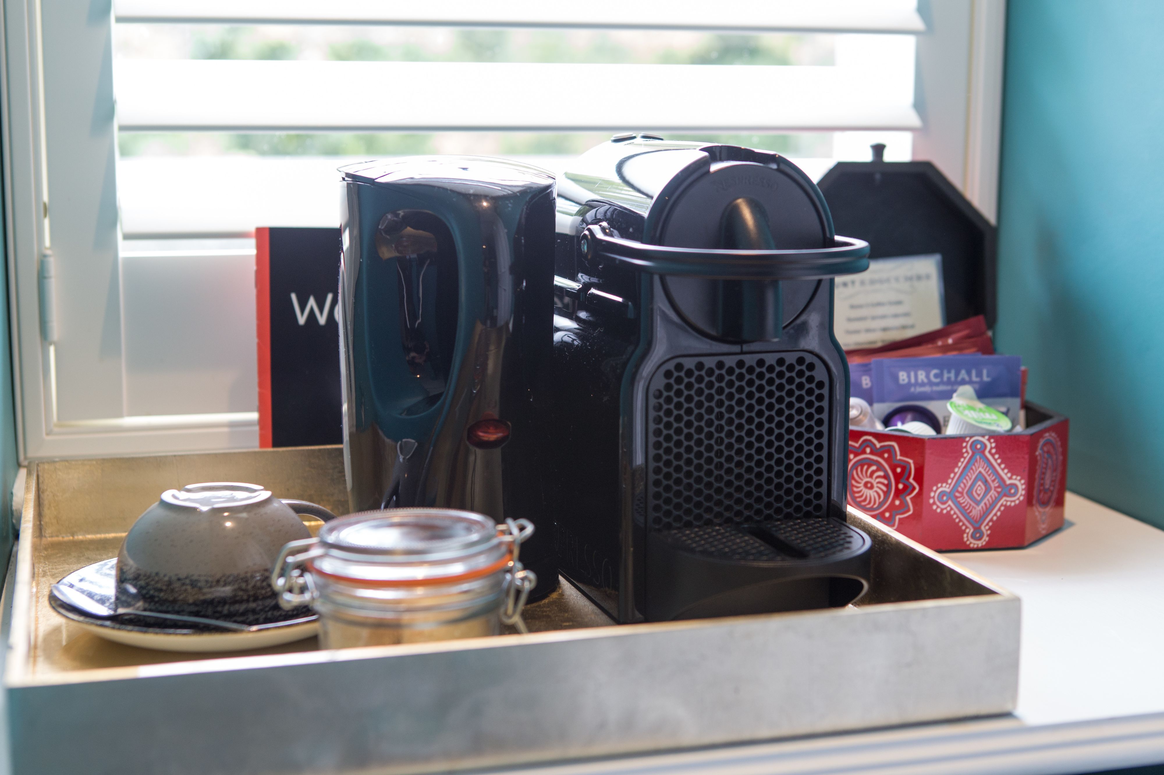 A coffee maker, a teapot, and a jar on a tray by a window.
