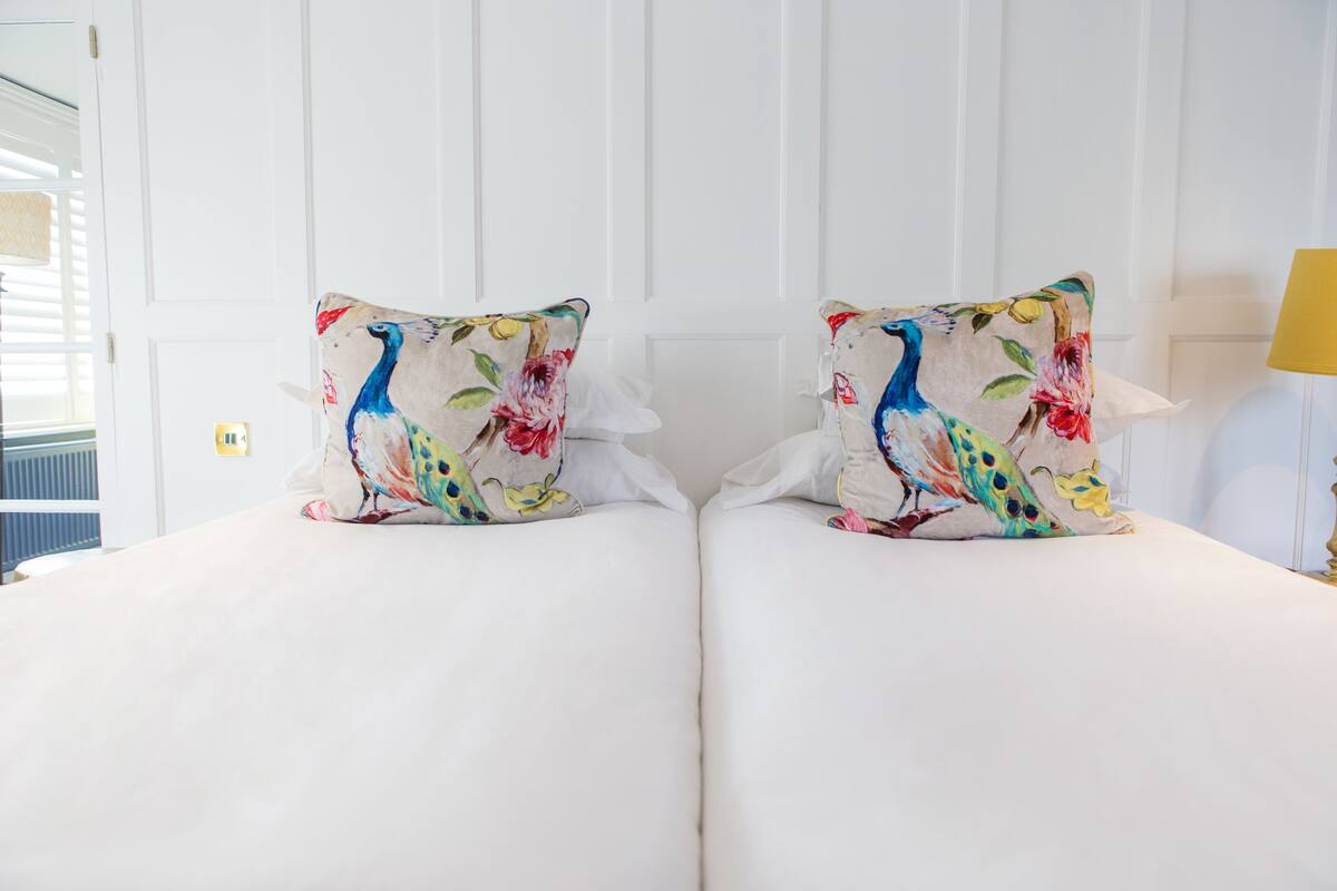Een tweepersoonsbed met witte beddengoed en twee decoratieve kussens met een pauwendessin.