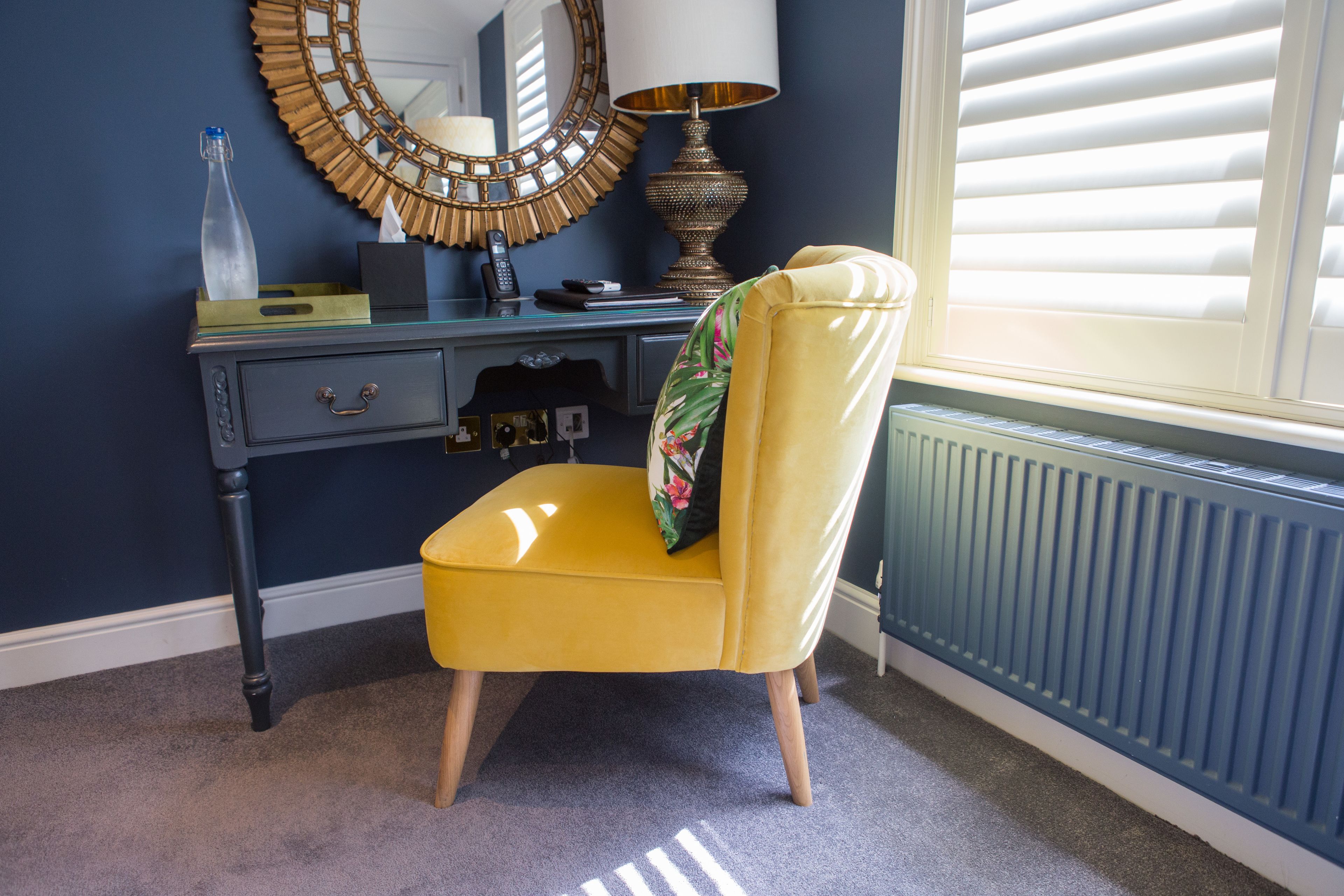 A room with a yellow chair, a desk with drawers, a mirror, a lamp, and a window with blinds.