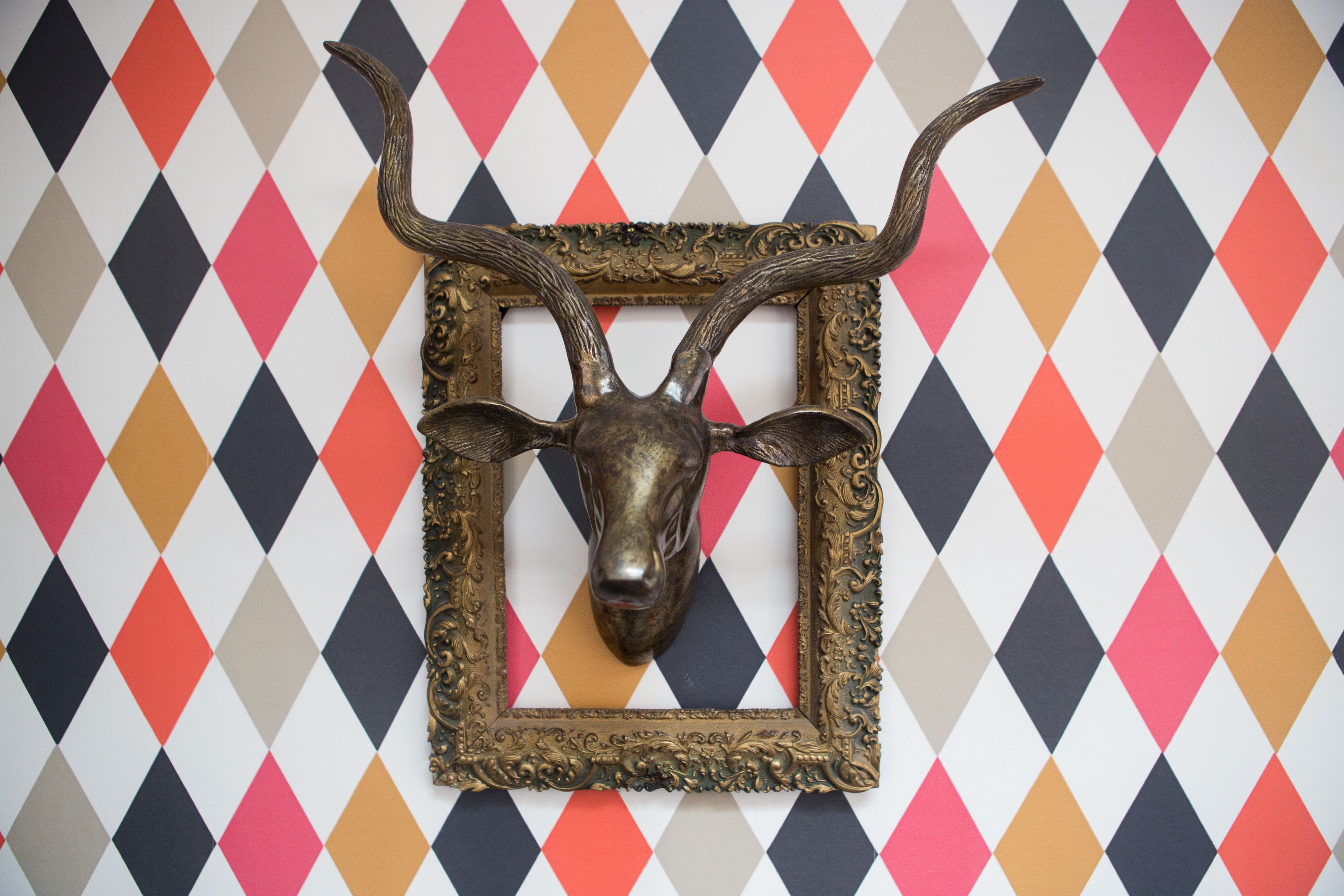Escultura de cabeça de animal fixada na parede, com chifres, emoldurada por um ornamento detalhado, em frente a uma parede com padrão de losangos.