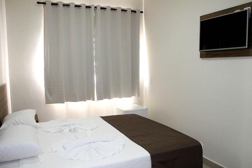 A hotel room with a bed, a bedside table, a TV mounted on the wall, and a window with curtains.
