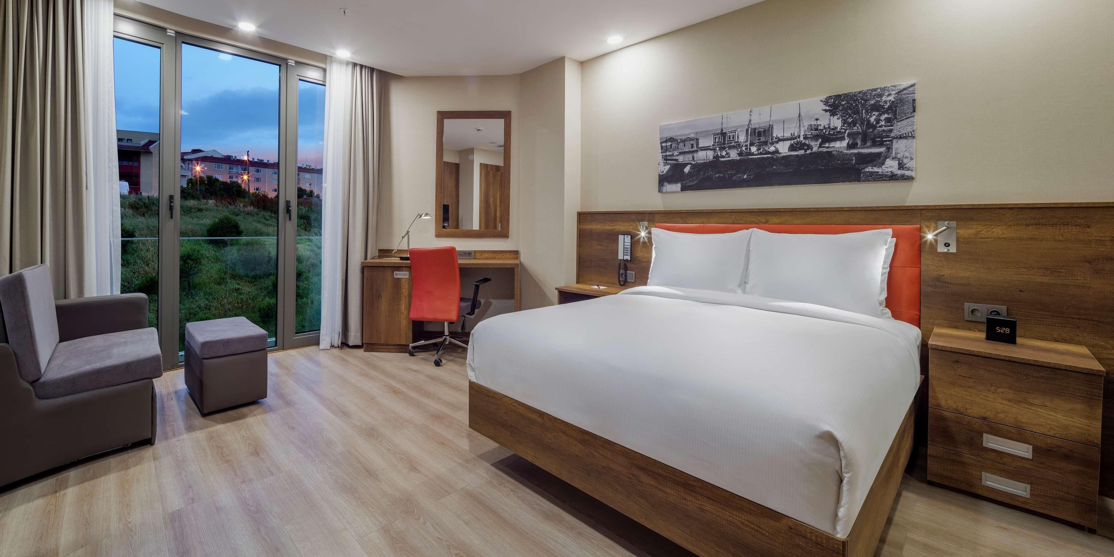 Une chambre d’hôtel avec un grand lit, un bureau avec une chaise, une table de chevet et une vue sur la verdure.