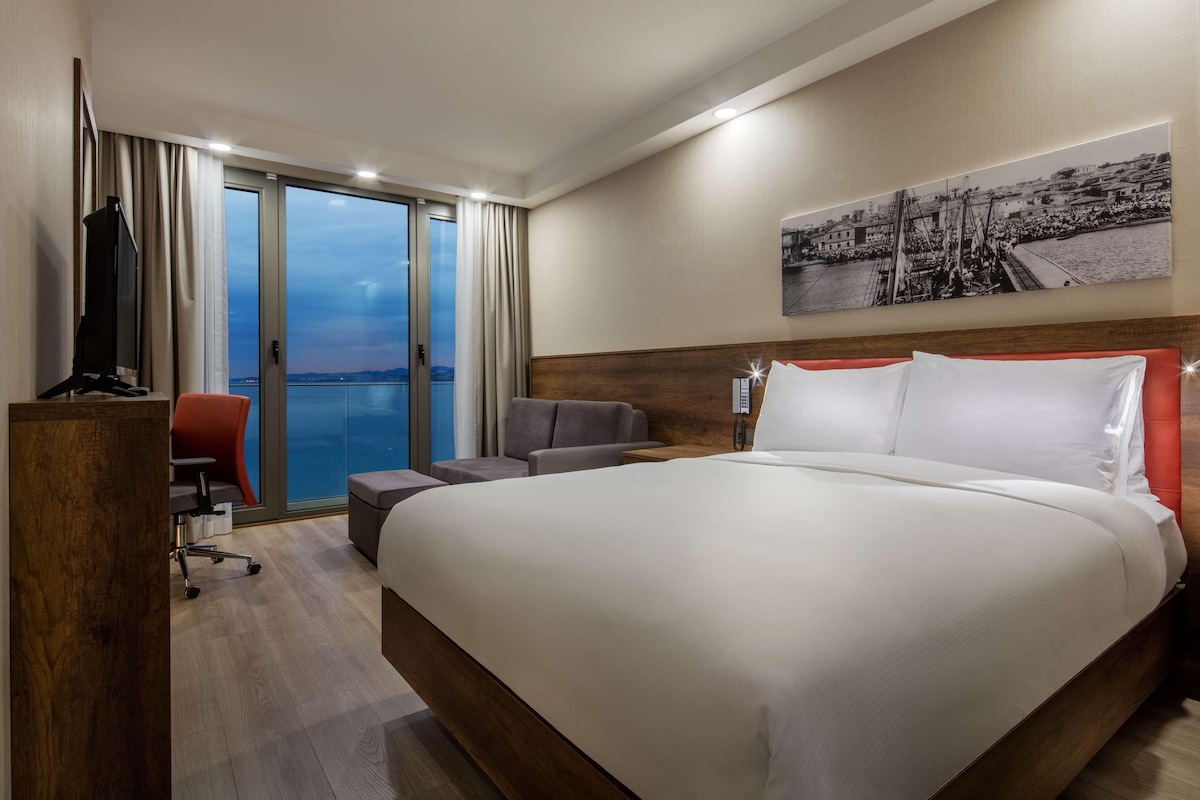 Une chambre d’hôtel avec un grand lit, un bureau avec une chaise, une télévision et une vue sur la mer, accessible par une porte coulissante.