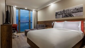 A hotel room with a large bed, a desk with a chair, a television, and a view of the sea through a sliding door. - Hampton by Hilton Canakkale Gallipoli (Gallipoli)