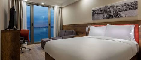 A hotel room with a large bed, a desk with a chair, a television, and a view of the sea through a sliding door.