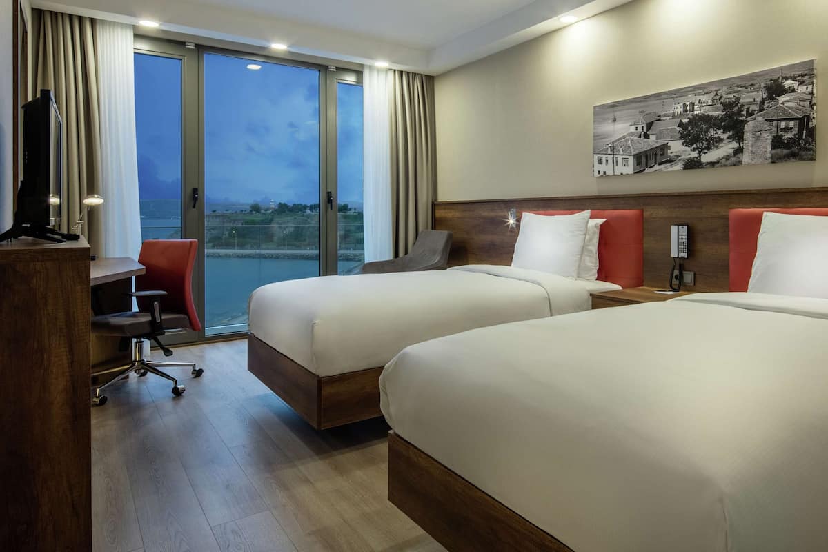 Habitación de hotel con dos camas, un escritorio, una silla y un ventanal con vistas a la ciudad.